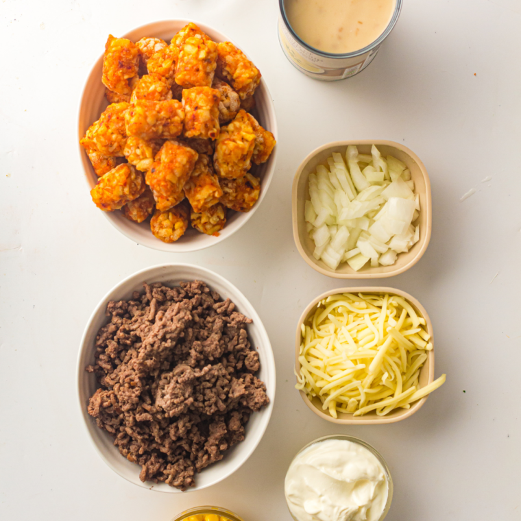 Ingredients for cowboy casserole including ground beef, corn, cream of mushroom soup, sour cream, cheddar, onion, and frozen tater tots