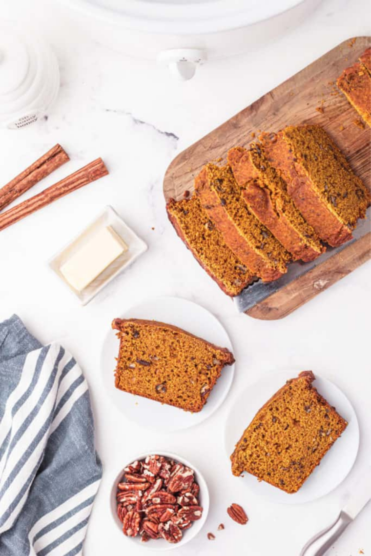 Overhead shot of sliced pumpkin bread on marble with pecans, butter, and cinnamon sticks