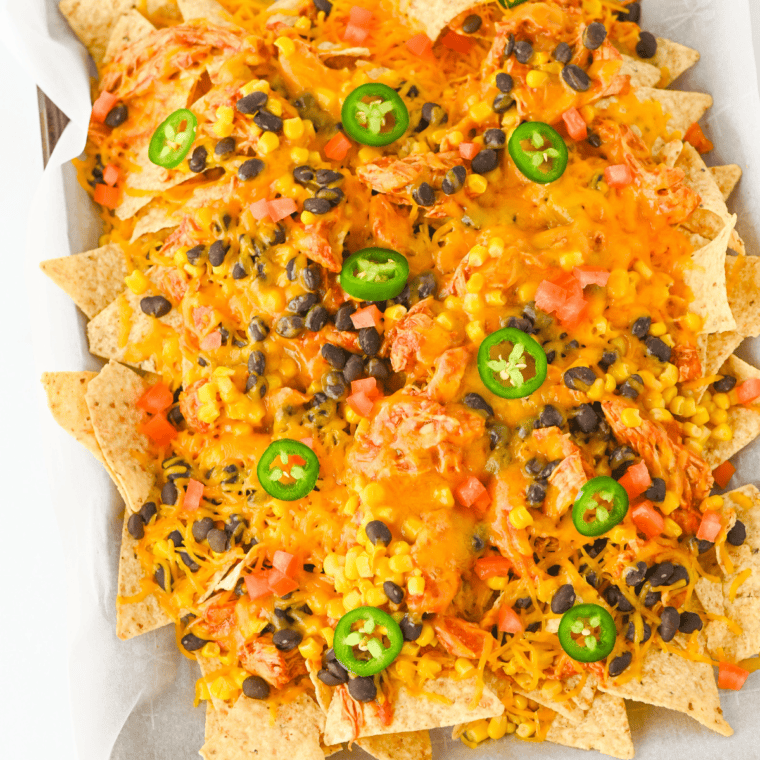 Crockpot chicken nachos on a parchment-lined sheet pan, loaded with shredded salsa chicken, melted cheddar, corn, black beans, diced tomatoes, and sliced jalapeños