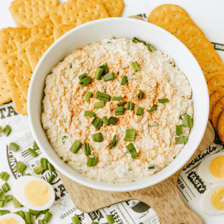 Deviled egg dip topped with paprika and green onions in a white serving bowl with Ritz crackers